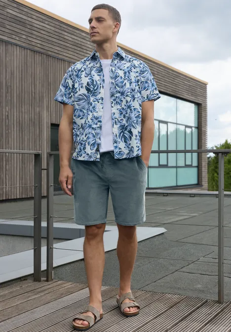 Homme sur terrasse de toit devant bâtiment bois-verre, porte chemise bleue à fleurs, short gris et sandales.