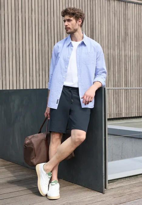 Homme décontracté en chemise ouverte, t-shirt, shorts, baskets blanches et sac en cuir devant un mur urbain