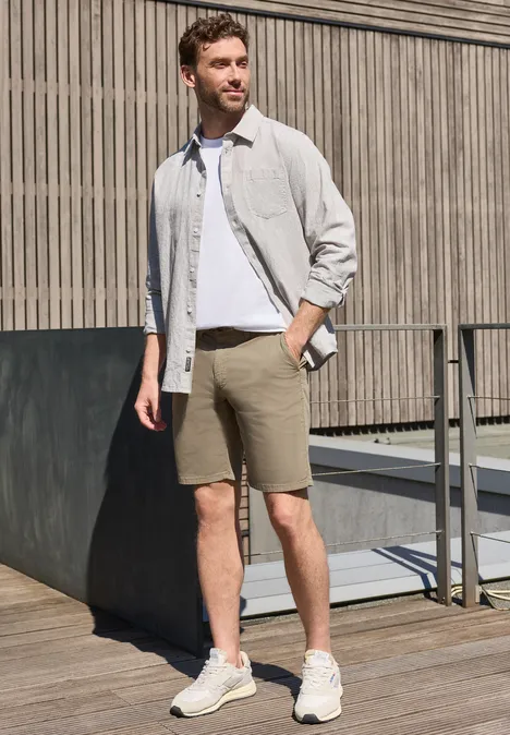 Ein Mann in weißem T-Shirt, hellgrauem Hemd, beigen Shorts und Sneakers steht auf einer Holzterrasse vor Holzpaneelen.
