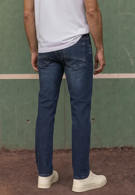 Ein Mann von hinten in blauen Jeans und weißen Sneakers, stehend vor einer grünen Wand.