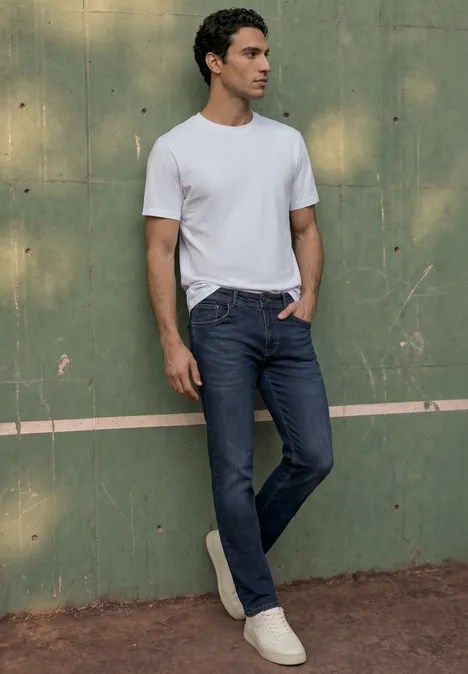Ein Mann trägt ein weißes T-Shirt und blaue Jeans, lehnt an einer grünen Wand.