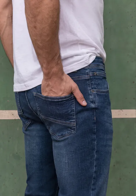 Mann von hinten in weißem T-Shirt und blauen Jeans lehnt an grünem Hintergrund, Hand in Gesäßtasche.