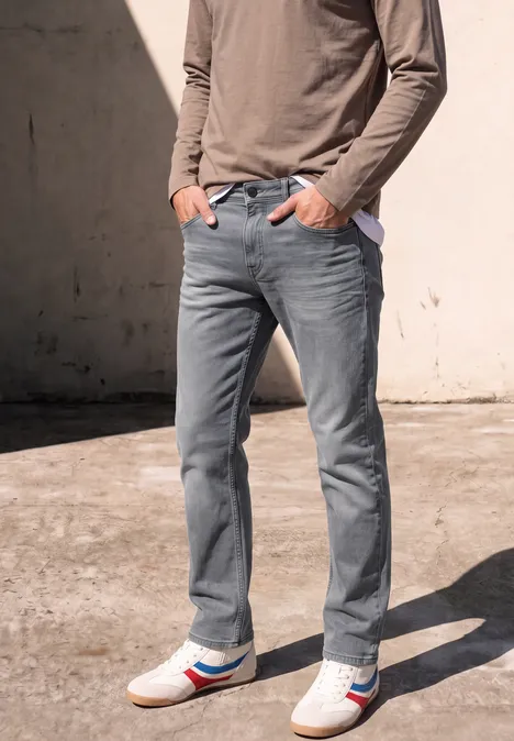 Straight Leg Jeans im Regular Fit mid grey random wash Mann trägt graue Jeans, langärmeliges Oberteil und weiße Sneaker mit roten und blauen Streifen vor Betonwand.