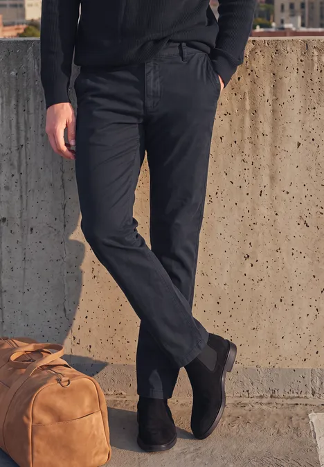 Person lehnt an Betonwand, trägt schwarze Hose und Stiefel, braune Ledertasche liegt daneben.