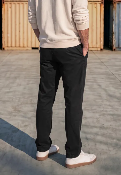 Slim Leg chino met flexibele tailleband basic black Achteraanzicht van iemand met zwarte broek en witte sneakers op een betonnen vloer voor containers.