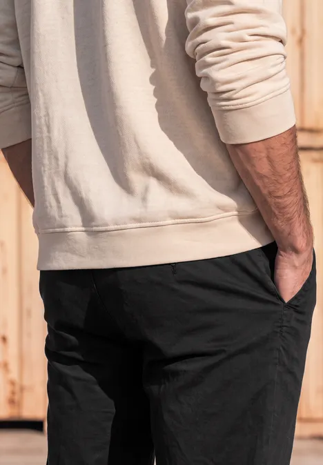 Slim Leg chino met flexibele tailleband basic black Een man in een beige trui en zwarte broek staat zijwaarts voor een houten wand, hand in de zak.