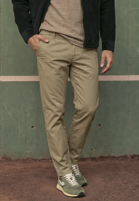 Een man staat voor een groene muur met beige chino’s en groene sneakers.