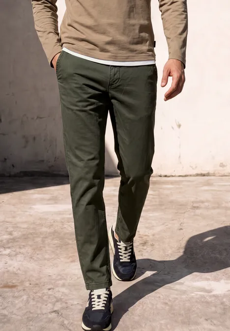Man in olijfgroene broek en sneakers loopt over een zonnige betonnen vloer.