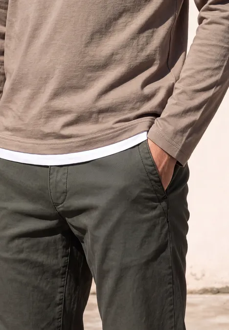Persoon in lichtbruine longsleeve over wit shirt en donkergroene broek, hand in de zak