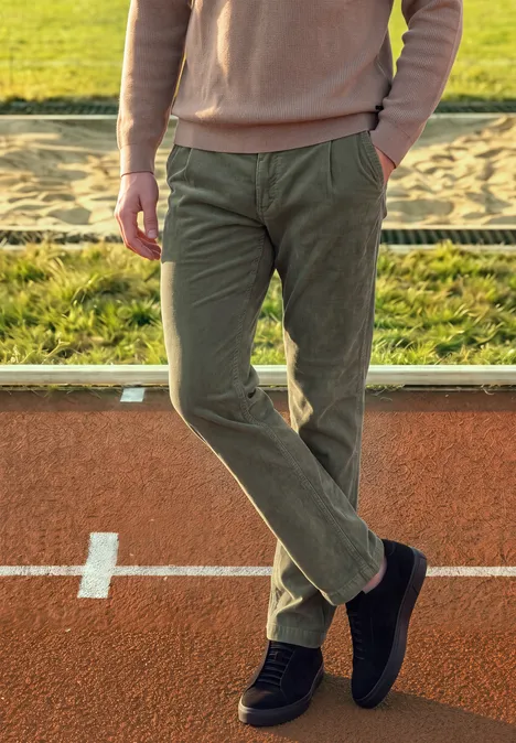 Une personne en pull rose, pantalon vert et chaussures noires se tient de manière décontractée sur une piste.