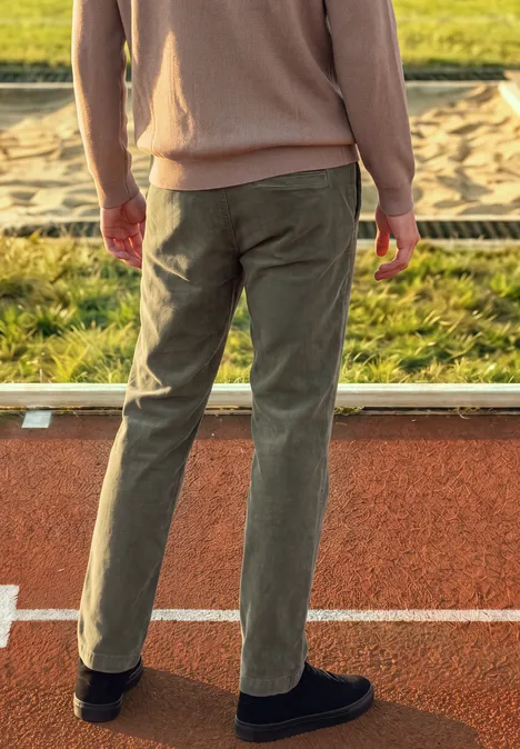 Une personne vue de dos en veste beige et pantalon vert olive se tient sur une piste en tartan près d'un bac à sable.
