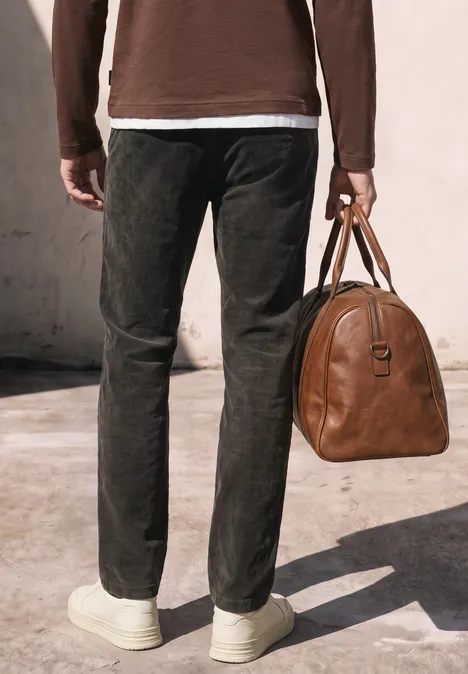 Pantalon chino en velours côtelé avec plis marqués au fer à repasser obsidian grey Homme en pantalon noir, baskets blanches, tenant un sac en cuir marron devant un mur clair.