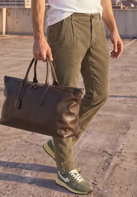 Pantalon chino en velours côtelé avec plis marqués au fer brass brown Homme en pantalon kaki et baskets porte un grand sac en cuir brun sur un toit.