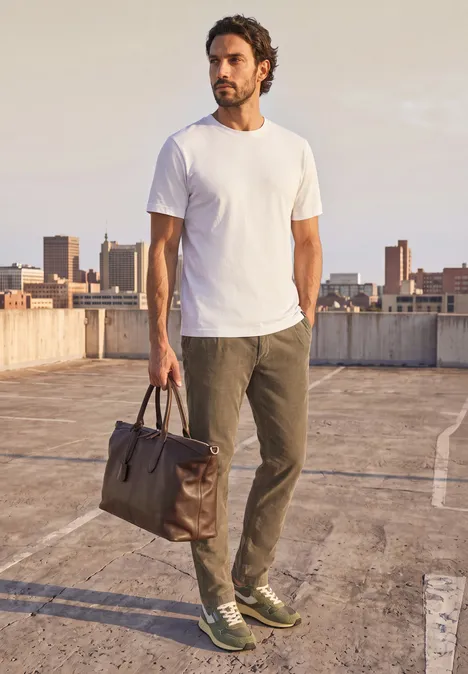 Pantalon chino en velours côtelé avec plis marqués au fer brass brown Un homme en t-shirt blanc et pantalon vert porte un sac sur un parking sur toit face à la skyline.