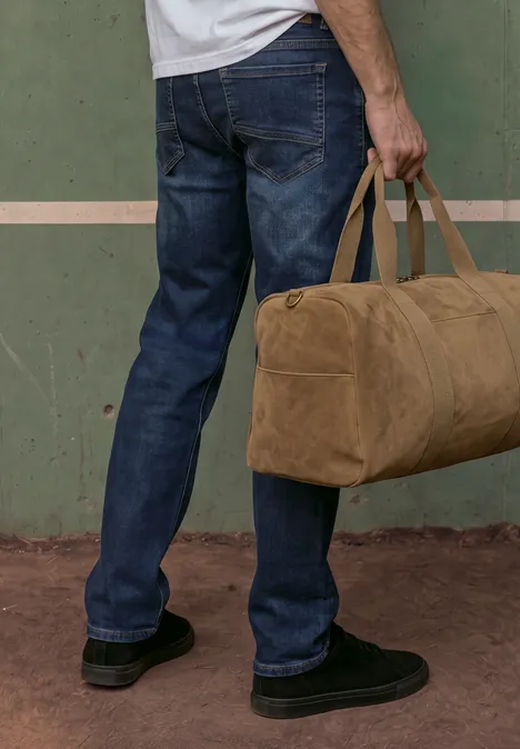 Mann trägt blaue Jeans und schwarze Schuhe, hält eine beige Reisetasche vor einer grünen Wand.