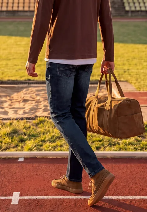 Jean Slim Leg à coupe ajustée indigo blue wash Un homme marche sur une piste d’athlétisme en tenant un sac de sport marron.