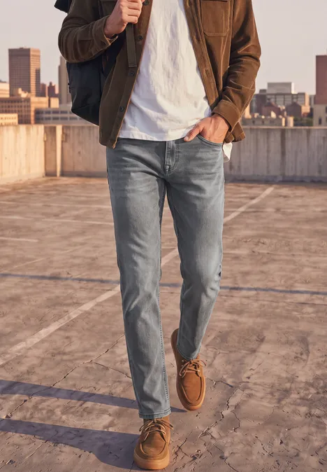 Slim Leg Jeans im Regular Fit mid grey random wash Eine Person geht auf einem Parkhausdach, trägt Jeans, weißes T-Shirt, braune Jacke und braune Boots.