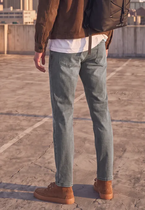 Slim Leg Jeans im Regular Fit mid grey random wash Person von hinten in Jeans und braunen Stiefeln mit Rucksack auf Dachparkplatz.