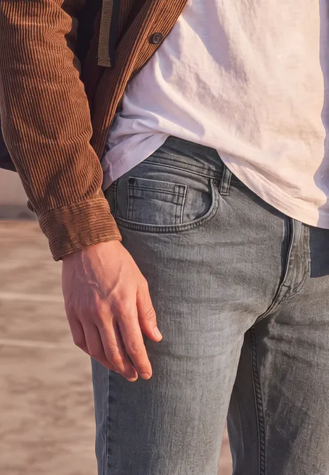 Slim Leg Jeans im Regular Fit mid grey random wash Nahaufnahme eines Oberkörpers in Jeans, weißem T-Shirt und brauner Jacke mit entspannter Hand.