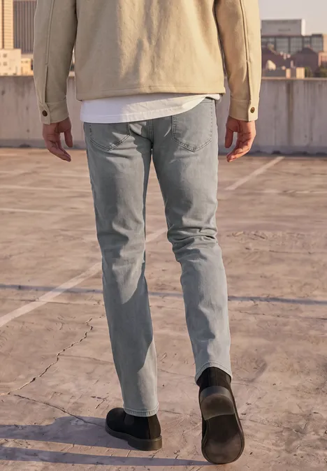 Slim Leg Jeans im Slim Fit mid grey random wash Ein Mann in Jeans, T-Shirt und Jacke geht auf einem leeren Dachparkdeck.