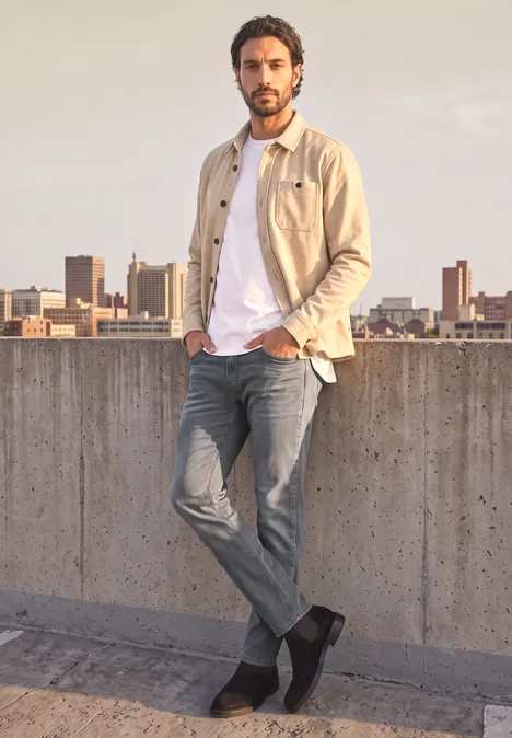 Slim Leg Jeans im Slim Fit mid grey random wash Mann mit Jeans und beiger Jacke lehnt an Betonmauer, Stadtsilhouette im Hintergrund.