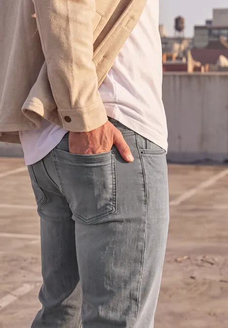 Slim Leg Jeans im Slim Fit mid grey random wash Mann steht auf Dach, trägt Jeans, weißes Shirt, beige Jacke, eine Hand in der Hosentasche.
