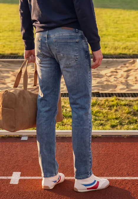 Mann in Jeans und Sneakern mit Reisetasche steht auf Laufbahn neben Sandgrube.