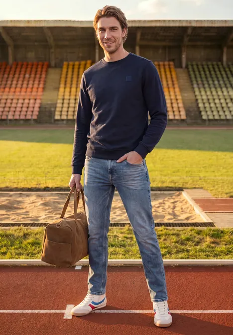 Ein Mann steht auf der Laufbahn vor dem Weitsprung-Sandgraben, hält eine Tasche, trägt Jeans und Pullover.