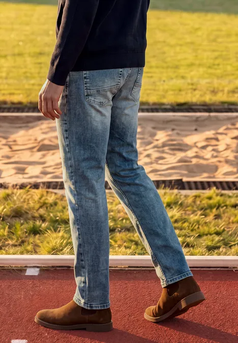 Eine Person in Jeans und braunen Schuhen geht auf einer Sportbahn neben einer Sandgrube.