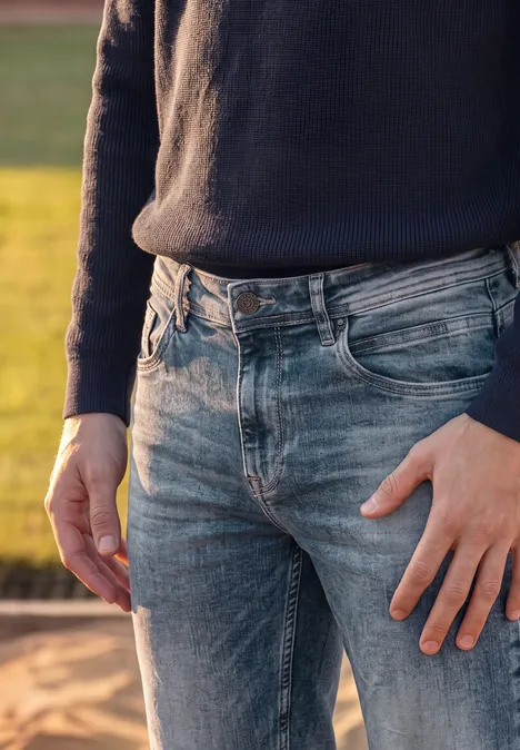 Eine Person trägt draußen blaue Jeans und einen dunklen Pullover und steht auf sandigem Untergrund.