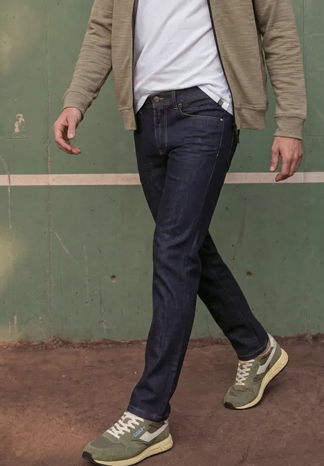 Een man loopt langs een groene muur, hij draagt donkere spijkerbroek en groene sneakers.