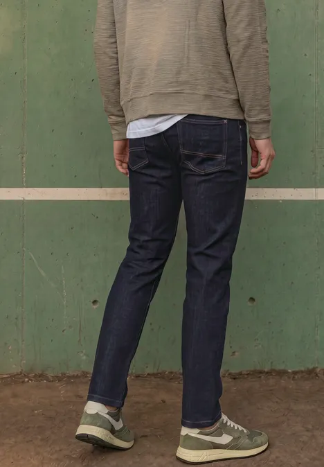 Persoon met rug naar camera draagt donkere jeans en sneakers voor een groene wand.