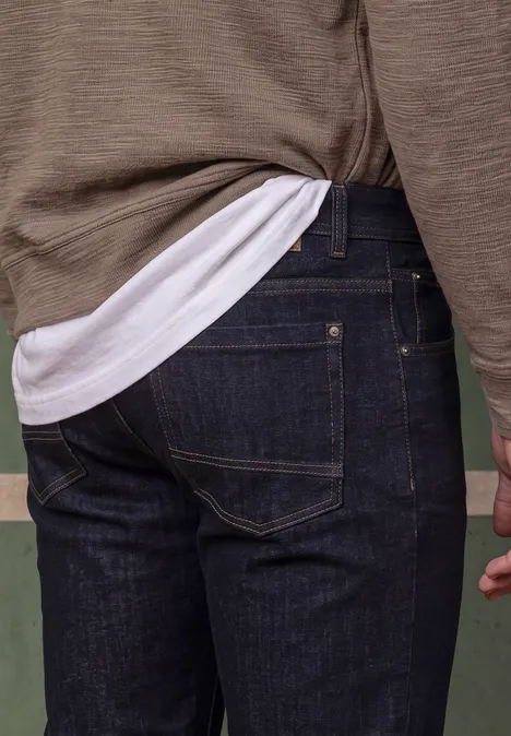 Achteraanzicht van een persoon in donkerblauwe jeans en groen shirt tegen effen achtergrond.