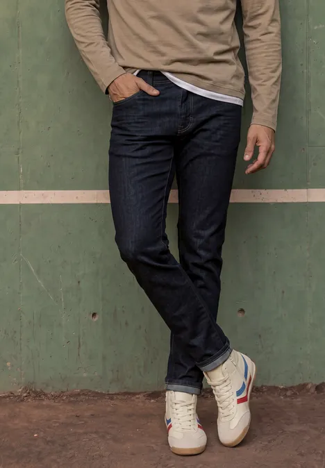 Slim Leg Jeans im Slim Fit dark blue rinsed wash Ein Mann in Jeans und Sneakers steht vor einer grünen Wand, eine Hand in der Hosentasche.