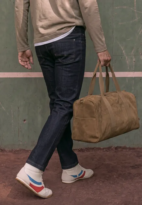 Slim Leg Jeans im Slim Fit dark blue rinsed wash Person in Jeans und Sneakern hält eine braune Reisetasche vor grüner Wand.