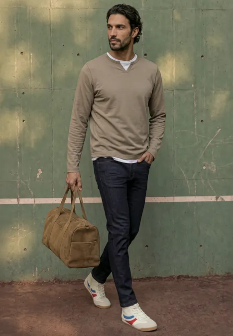 Slim Leg Jeans im Slim Fit dark blue rinsed wash Ein bärtiger Mann in beigem Pulli, dunkler Hose und Sneakern trägt eine Tasche vor einer grünen Wand.