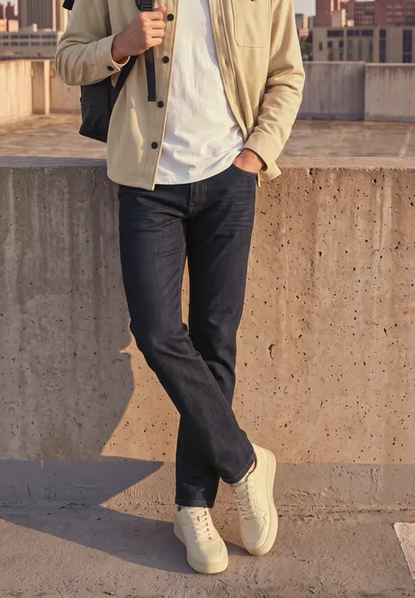 Straight Leg Jeans im Regular Fit dark blue rinsed wash Ein Mann in dunklen Jeans, beigem Mantel und weißen Sneakern steht lässig vor einer Betonmauer, urbane Kulisse.