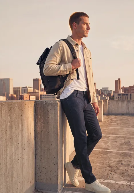 Straight Leg Jeans im Regular Fit dark blue rinsed wash Ein Mann mit Rucksack lehnt auf einem Parkdeck, im Hintergrund ist eine Stadtlandschaft zu sehen.
