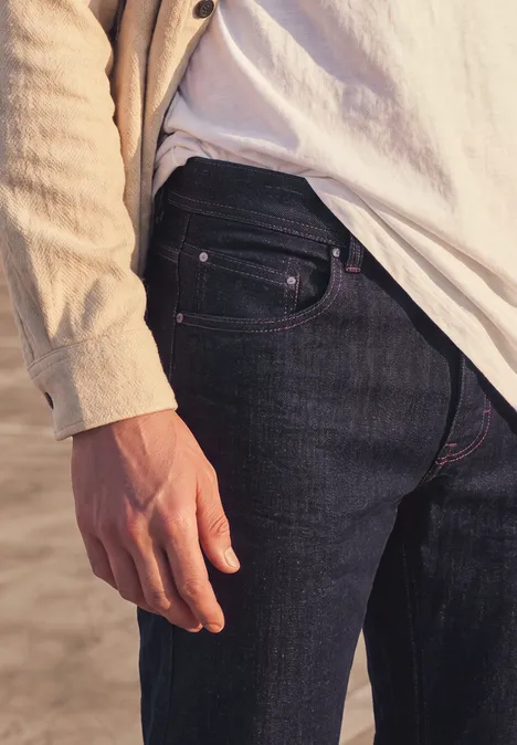Straight Leg Jeans im Regular Fit dark blue rinsed wash Seitliche Nahaufnahme einer Person am Strand in dunkler Jeans, weißem T-Shirt und beiger Jacke.
