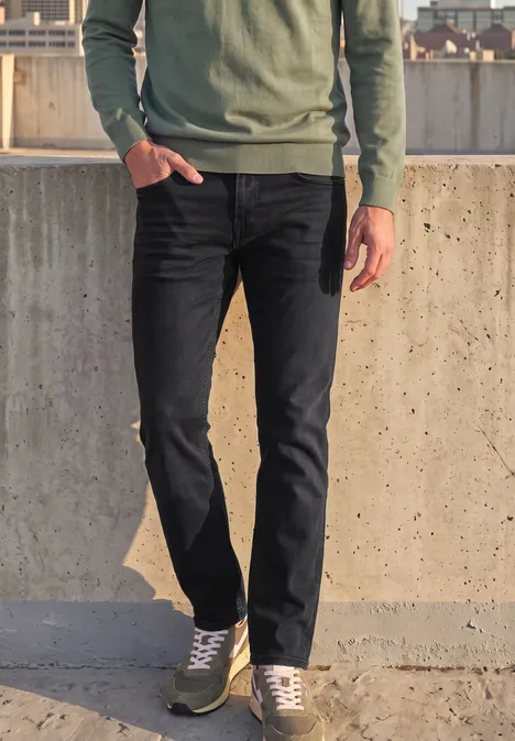 Eine Person in schwarzer Jeans, grauen Sneakern und grünem Pullover lehnt an einer Betonmauer im Freien.