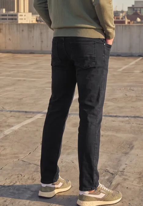 Ein Mann von hinten in schwarzer Jeans, olivgrünem Sweatshirt und grünen Sneakern auf einem Parkdeck.