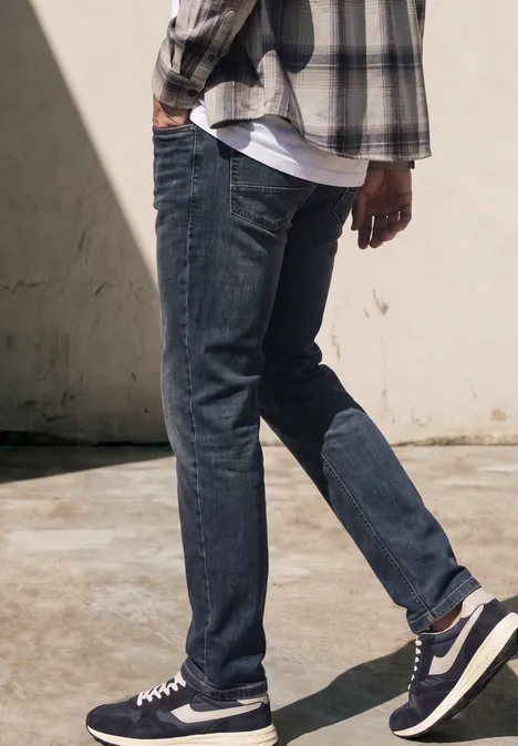 Untere Körperhälfte einer Person in blauen Jeans, Sneakern und kariertem Hemd vor heller Wand auf Betonboden.