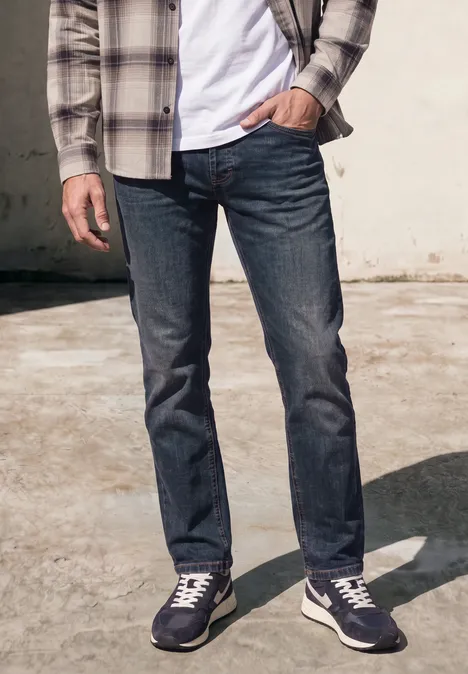 Mann im Freien, trägt dunkelblaue Jeans, weiße Sneakers, weißes T-Shirt und kariertes Hemd.