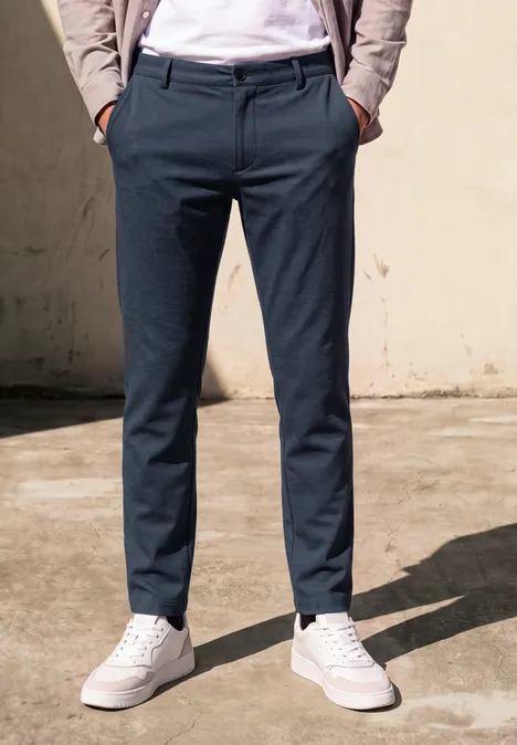 Een persoon draagt een donkerblauwe broek en witte sneakers voor een lichte muur.