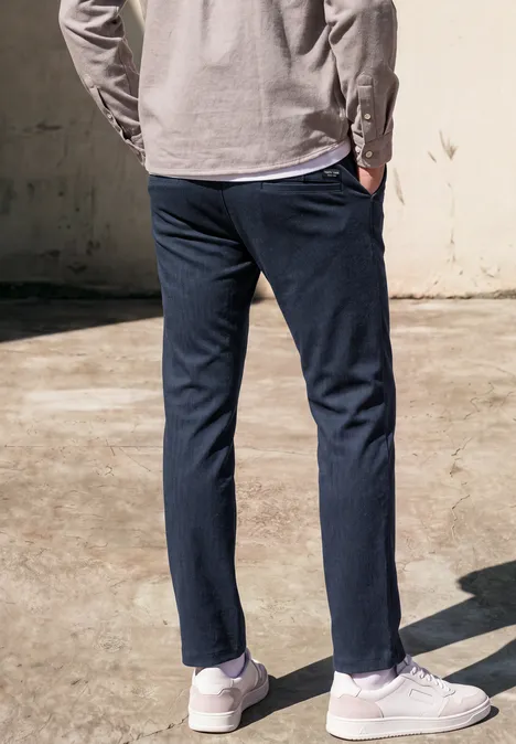 Persoon van achteren in donkerblauwe broek, grijs shirt en witte sneakers op zonnige betonnen vloer.