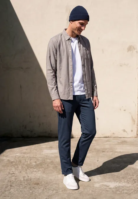Man met muts, open overhemd over t-shirt, slim-fit broek en witte sneakers, staat ontspannen in zonlicht.
