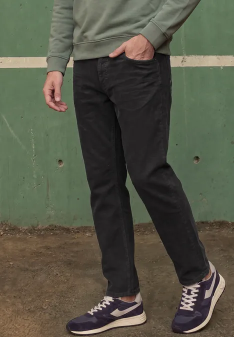 Eine Person trägt ein grünes Oberteil, schwarze Jeans und blaue Sneaker vor einer grünen Wand.