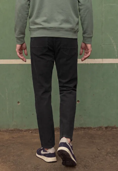 Person von hinten, trägt schwarze Jeans und blaue Sneaker, steht vor einer grünen Wand.