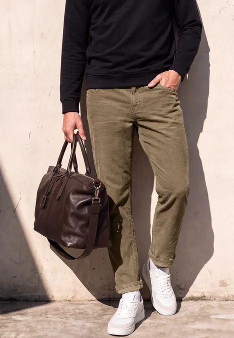 Mann in olivgrünen Hosen, weißen Sneakern und schwarzem Pullover hält braune Ledertasche vor heller Wand.
