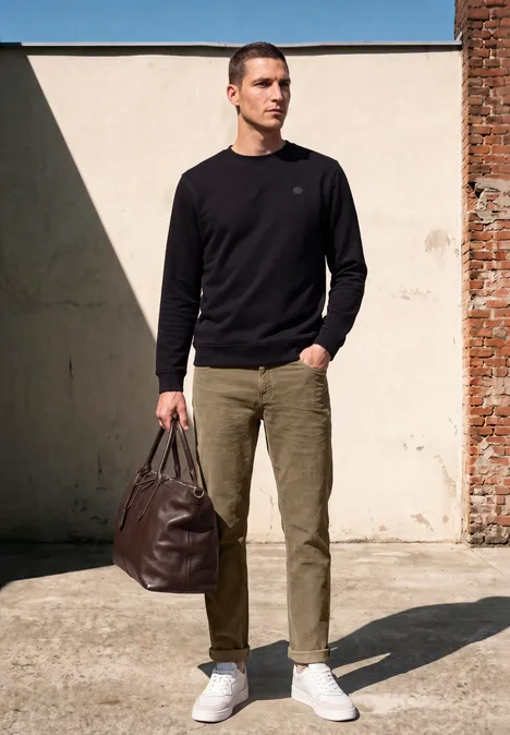 Ein Mann in schwarzem Pullover, grünen Hosen und weißen Sneakern trägt braune Tasche.